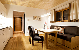 Wohnküche mit Holzbank, Esstisch, zwei Lederstühlen, Küchenzeile, Fenster mit Vorhang und Bild an weißer Wand.