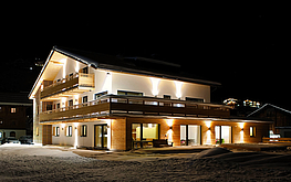 Nachtaufnahme von Das Tannberg: beleuchtetes Holz-Weiß-Haus mit Balkonen und verschneitem Vorplatz.