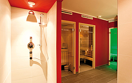 Wellnessbereich mit offener Dusche, roter Wand, Fliesenboden, Saunatüren und grün beleuchtetem Ruheraum.