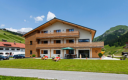 Ferienhaus „Das Tannberg“ mit Holzfassade und gepflegtem Garten in Lech am Arlberg, umgeben von grünen Hügeln und blauen Himmel.