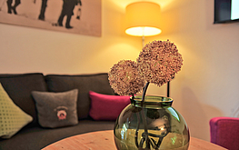 Nahaufnahme einer Blumenvase mit Trockenblumen auf einem Tisch vor einem Sofa mit bunten Kissen und gemütlichem Ambiente.