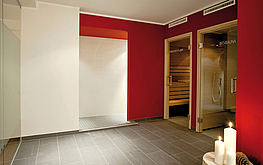Wellnessbereich mit dunklem Fliesenboden, roter Wand, zwei Saunatüren, Kerzen auf weißem Podest